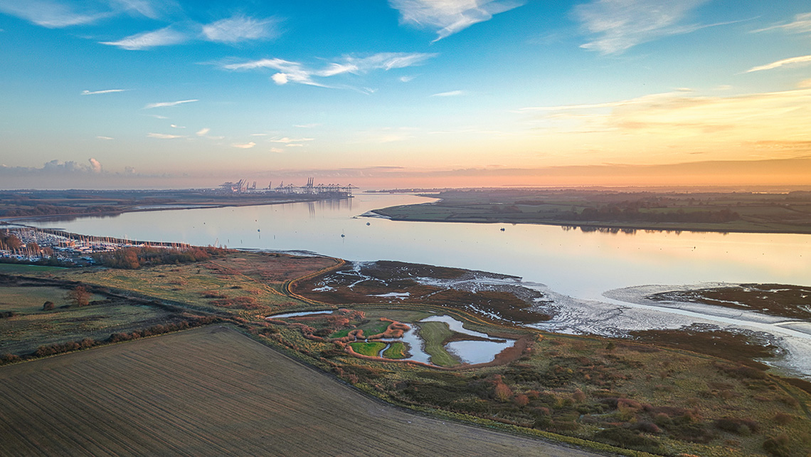 A river landscape at sunset with fields and wetlands in the foreground and distant industrial structures on the horizon.