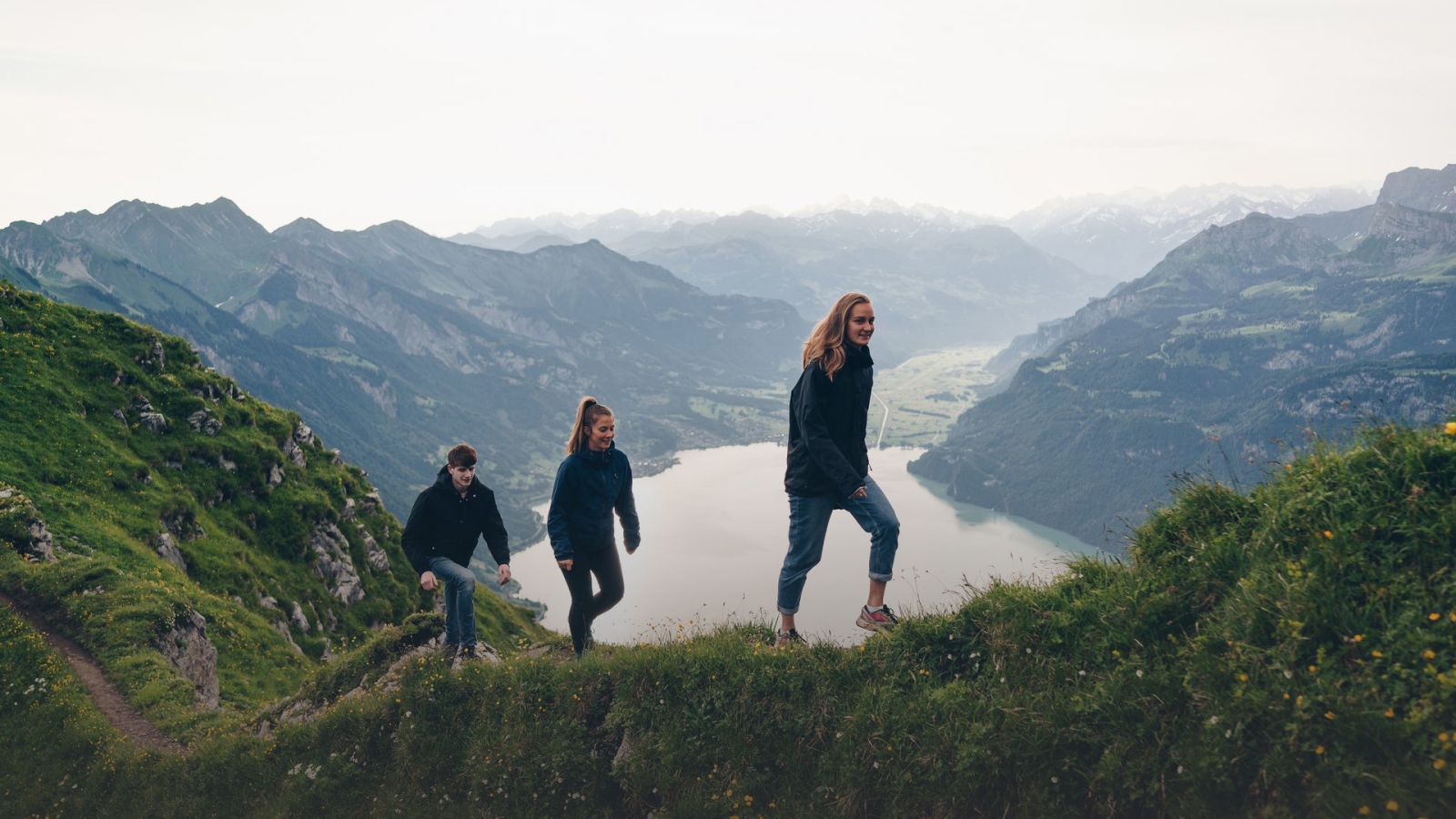 Group of friends hiking on a mountain trail with scenic lake and alpine peaks in the background.